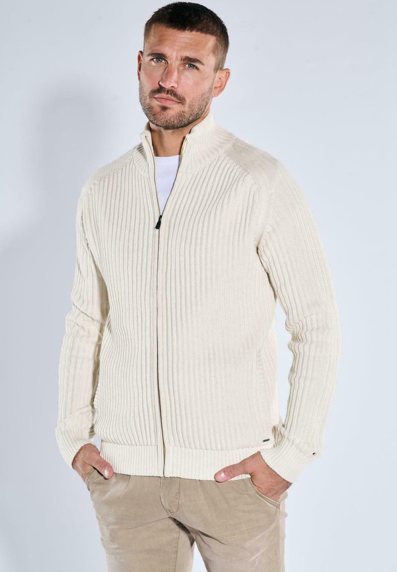 Mann mit kurzem Haar und Bart, der einen cremefarbenen gerippten Pullover mit Reißverschluss und beigen Hosen trägt, steht mit den Händen in den Taschen vor einem schlichten Hintergrund.