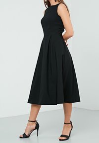 Černé bezramínkové šaty s přiléhavým korzetem a volnou sukní, vyrobené z hladké látky. Kombinované s černými sandály na vysokém podpatku se středními pásky.