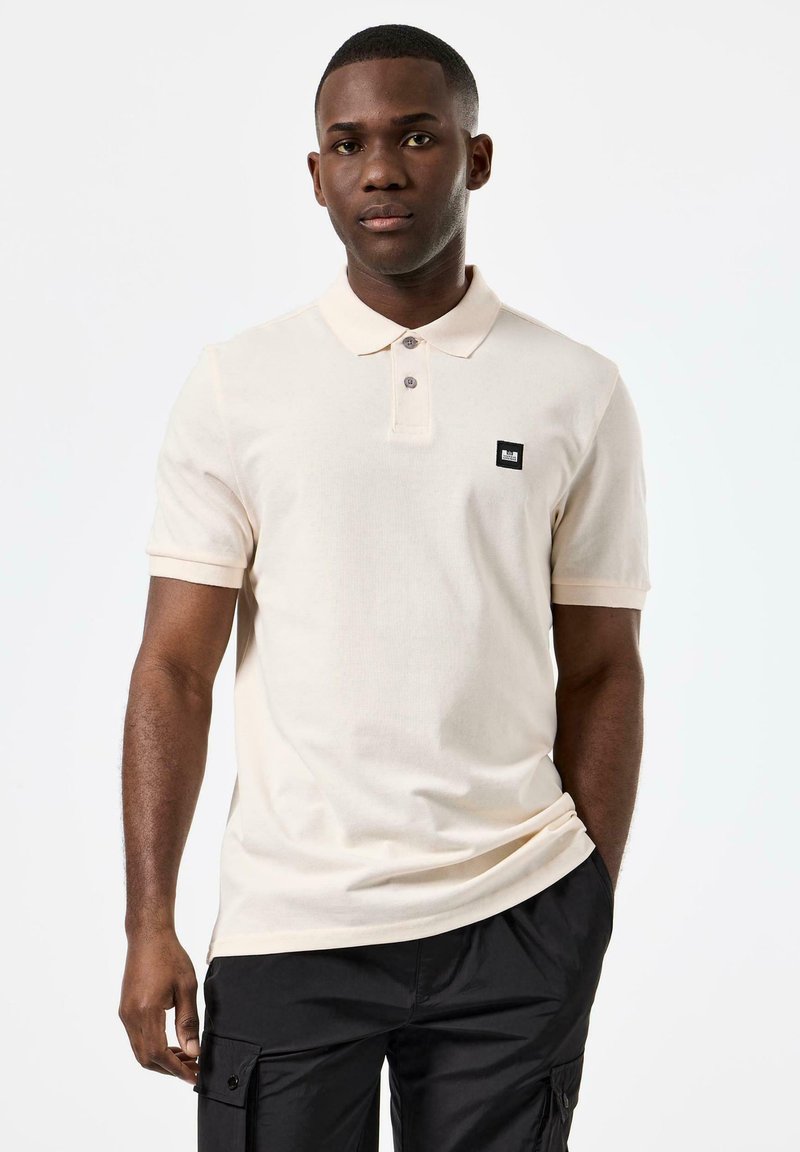 Beige pikéskjorte med korte ermer, to-knapp placket, og en svart logo-patch. Brukt med svarte cargo-shorts, enkel bakgrunn.