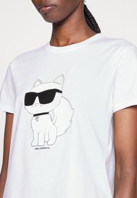Tricou alb din bumbac, cu un desen de pisică în stil cartoon purtând ochelari de soare și zgardă, conturată în negru, cu textul "KARL LAGERFELD" sub design.