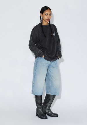 Camicia a maniche lunghe oversize grigio carbone, jeans a gamba larga azzurri, stivali in pelle neri al ginocchio, capelli intrecciati, in piedi su uno sfondo chiaro.