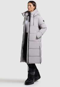 Ljusgrå, vadderad lång kappa med hög krage, fickor med dragkedjor och justerbara dragsnören; bärs med svarta vinterstövlar.