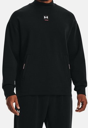 Mann trägt schwarzen Langarm-Pullover mit Kängurutasche und Under Armour-Logo auf der Brust sowie auf der Hosentasche vor einfachem Hintergrund.