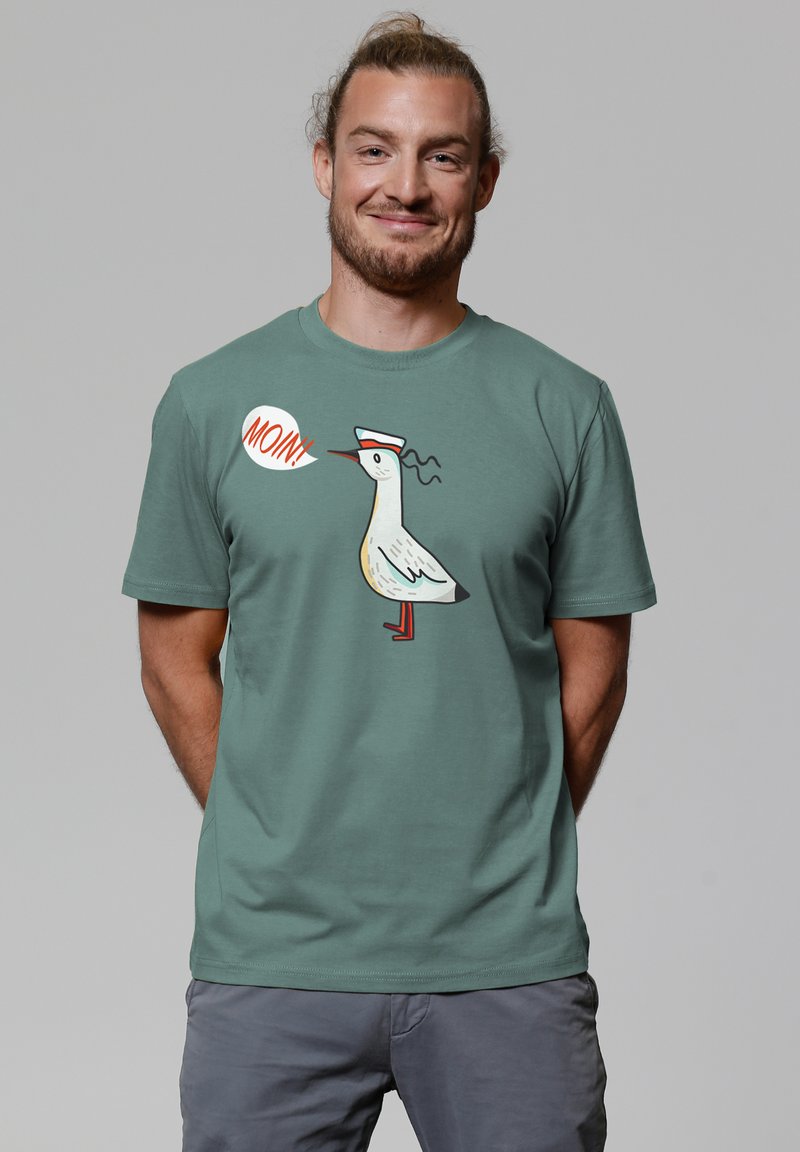 Baumwoll-T-Shirt in gedämpftem Grün mit einem Cartoon-Möwe im Matrosenhut und einer Sprechblase, die "Moin!" sagt. Bedrucktes Design in leuchtenden Farben.