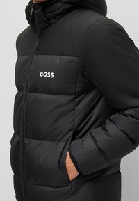 Svart puffjacka med kviltat mönster, logotyp på bröstet och dragkedja framtill. Inkluderar huva och elastiska ärmslut.