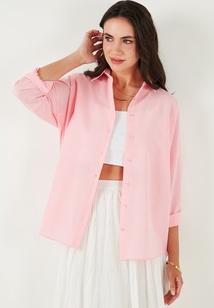 Light pink button-up shirt made of soft fabric, featuring a relaxed fit, roll-up cuffs, and a classic collar. Smooth texture and minimal detailing.