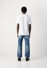 Person mit dunkler Hautfarbe und kurzen Dreads, die ein schlichtes weißes Kurzarmhemd, ausgebleichte blaue Jeans und schwarze Schuhe trägt und vor einer weißen Wand steht.