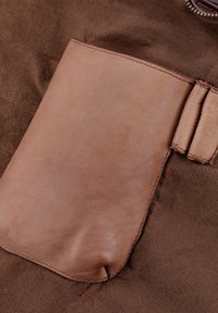 Brauner Stoff mit einer großen, strukturierten Tasche aus braunem Leder. Die Tasche hat sichtbare Nähte und eine glatte Oberfläche, die im Kontrast zum Stoff steht.