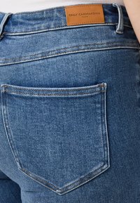 Μπλε τζιν παντελόνι με σχέδιο ορθογώνιας τσέπης, δερμάτινη ετικέτα στη μέση και λεπτομέρειες αντίθετης ραφής.