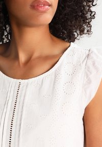 Blouse en coton blanc avec broderie ajourée, dotée d'un col rond, de manches courtes froncées et de détails de pinces verticales.