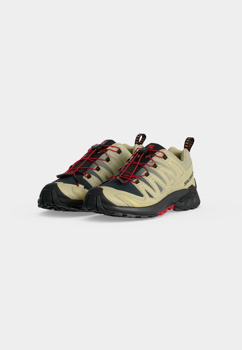 Paire de chaussures de trail beiges et noires avec lacets élastiques rouges et semelles noires robustes sur fond gris.