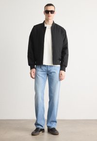 Svart bomberjacka över en vit skjorta, matchat med ljusblå jeans och svarta sneakers. Modellen bär mörka solglasögon. Enkel, avslappnad design.