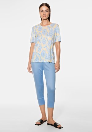 Helles blaues kurzärmliges Oberteil mit gelbem und blauem Paisleymuster, kombiniert mit hellblauen Cropped-Hosen und schwarzen Rutsch-Sandalen.