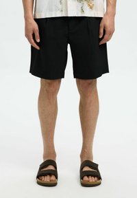 Černé kraťasy s rovnou siluetou a jemnými záhyby, doplněné tmavými sandály. Tkanina se zdá být lehká a s hladkým povrchem.