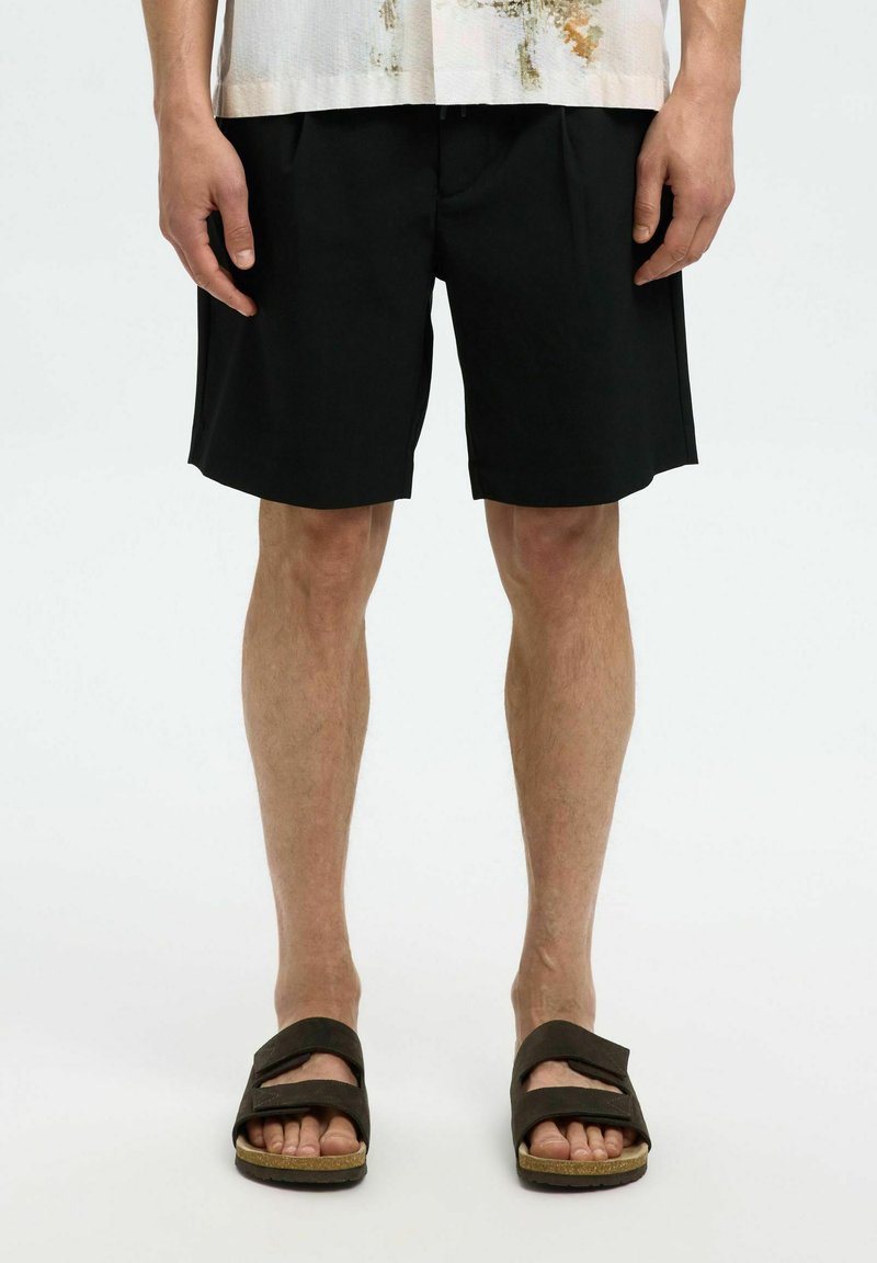 Černé kraťasy s rovnou siluetou a jemnými záhyby, doplněné tmavými sandály. Tkanina se zdá být lehká a s hladkým povrchem.