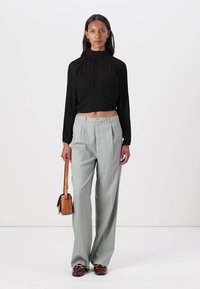 Top lungo a maniche lunghe plissettato nero abbinato a pantaloni a vita alta grigio chiaro e scarpe marroni, con una piccola borsa a tracolla color beige.