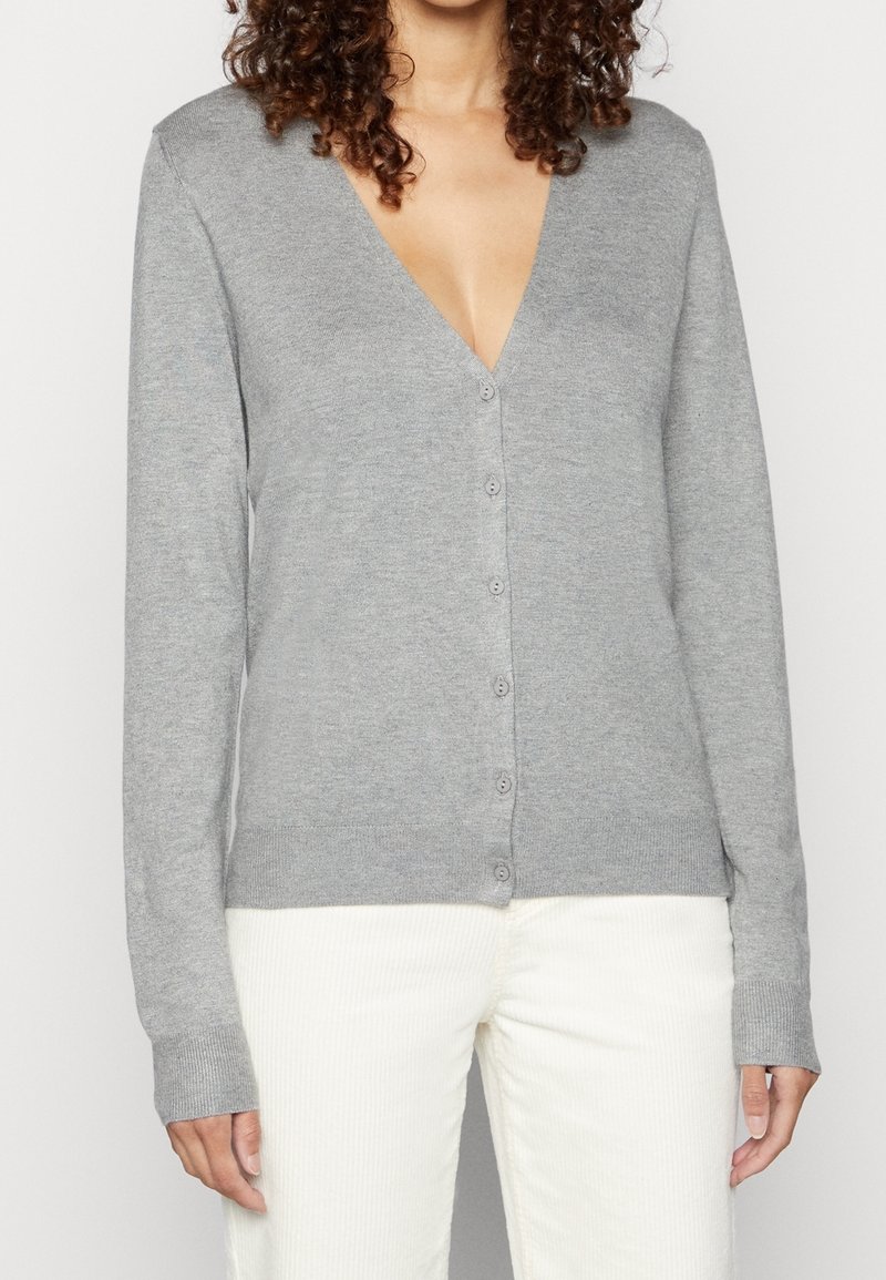 Femme portant un cardigan gris clair boutonné à manches longues et un pantalon blanc, montrée du cou aux hanches sur un fond uni.