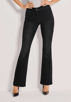 Schwarze Slim-Fit-Jeans mit ausgestellten Beinen, kombiniert mit schwarzen High Heels und einem grauen Oberteil, Hände auf den Hüften ruhend.