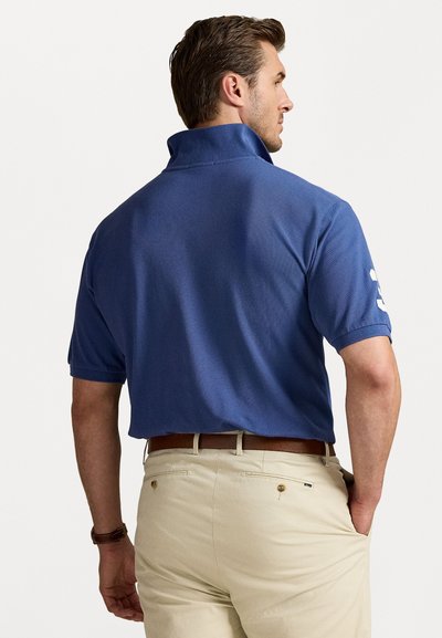 Tricou polo de culoare bleu marin cu guler ridicat, mâneci scurte și un accent alb cu numărul 3. Puratat cu pantaloni bej deschis și o curea maro.