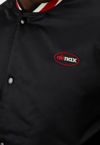 Veste en satin noire avec boutons-pression et finition du col rouge, blanc et noir, arborant un logo brodé "aii max" en rouge et blanc.