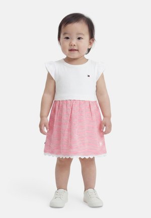 Niña pequeña con una camiseta blanca de mangas acampanadas y una falda a rayas rosas y blancas, de pie y con zapatillas blancas sobre un fondo blanco.