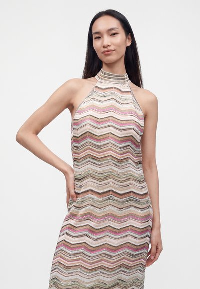 Jeune femme aux longs cheveux foncés portant une robe sans manches à col haut avec des motifs en zigzag multicolores, debout avec une main sur la hanche.
