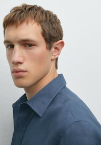 Blue button-up shirt with a collar, featuring a soft texture and smooth fabric. Model has short, light brown hair and neutral expression.