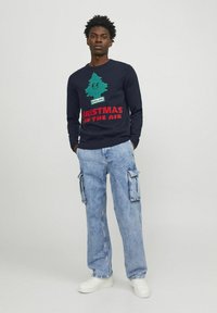 Marineblauer Pullover mit grafischem grünen Weihnachtsbaum und kräftigem roten Text. Kombiniert mit hellblauen Cargohosen mit großen Seitentaschen.