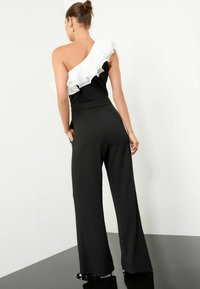 Zwarte jumpsuit met wijde broekspijpen, met een één-schouder ontwerp en een witte ruffle accent. Soepele stof met een aansluitende buste.