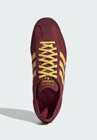 Bordeaux sneakers med gule snørebånd og tre sidelister. Har en tekstureret hæl og en mærket tunge. Blødt stofoverdel og gummisål.