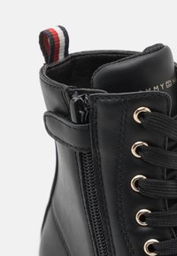 Stivale alla caviglia in pelle nera con zip laterale, occhielli dorati, lacci neri e una linguetta in tessuto a righe nella parte superiore. Texture liscia in tutto lo stivale.
