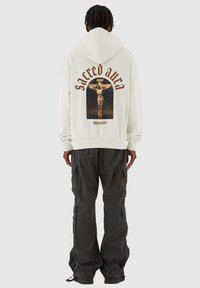 Weißer Hoodie mit einem großen Grafikdesign einer Kreuzigung und dem Text "sacred aura" in Braun, kombiniert mit dunklen Cargo-Hosen.