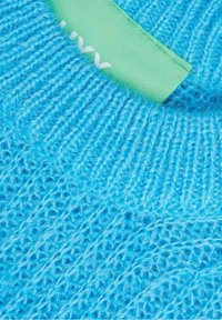 Tissu tricoté bleu vif avec un col côtelé et une étiquette de vêtement verte partiellement visible avec du texte blanc.