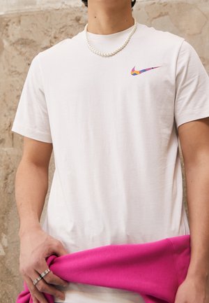 Personne portant un t-shirt blanc Nike avec un logo swoosh multicolore, tenant un vêtement rose vif et portant un collier de perles ainsi que des bagues.