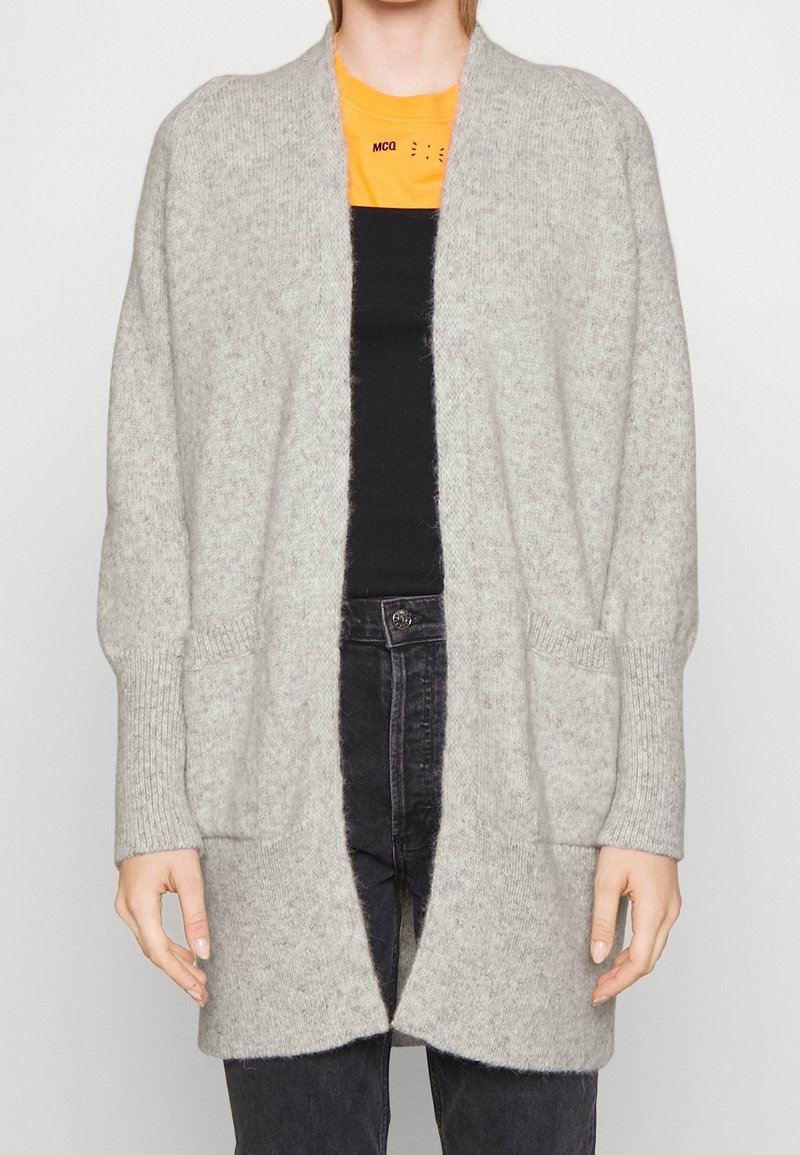 Light gray knitted cardigan with open front, ribbed cuffs, and two front pockets. Worn over a black top and dark jeans. Soft texture.