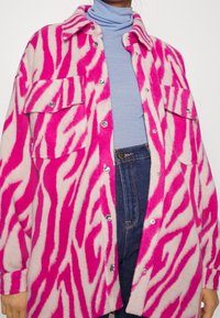 Överdimensionerad jacka med rosa och krämfärgad zebramönster i mjukt tyg med silverknappar och två framfickor, bärs över en ljusblå polotröja.
