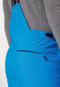 Gros plan d'une personne portant un pantalon à bavette bleu vif avec une poche arrière zippée et une taille ajustable par-dessus un haut gris à manches longues.