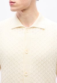 Vīrietis valkā krēmkrāsas adītu kardiganu ar teksturētu rakstu un trim redzamām pogām, apkakle nolocīta uz leju.