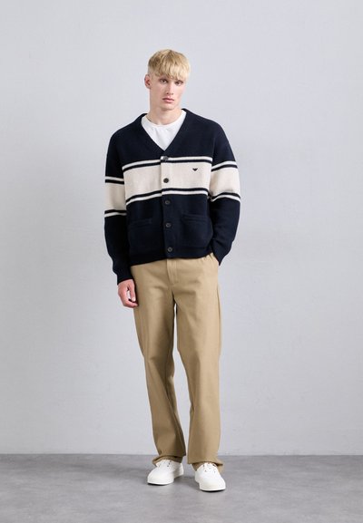 Ναυτικό πλεκτό ζακέτα με κρέμα ρίγες, κλείσιμο με κουμπιά και τσέπες, συνδυασμένη με μπεζ παντελόνια και λευκά αθλητικά παπούτσια, σε ουδέτερο φόντο.