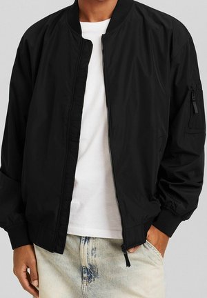 Mann trägt eine schwarze Bomberjacke mit Reißverschluss über einem weißen T-Shirt und hellblauen Jeans, Hände entspannt an den Seiten in lockerer Pose.
