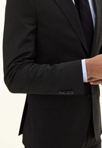 Zwarte blazer met een gestructureerde afwerking en een vierknopen manchetdetail, over een lichtblauw overhemd en een donkere das, gedeeltelijk zichtbaar.