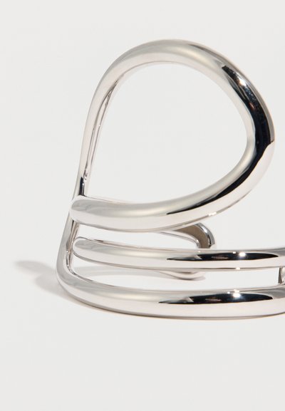 Λαμπερό ασημένιο βραχιόλι με ανοιχτό σχέδιο που παρουσιάζει επικαλυπτόμενες καμπυλωτές λωρίδες, σε απλό λευκό φόντο.