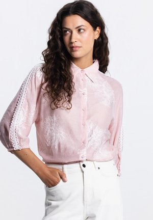 Vrouw met lang krullend haar, gekleed in een lichtroze blouse met knopen en witte bloemenborduursels, witte broek, hand in de zak, kijkt opzij.