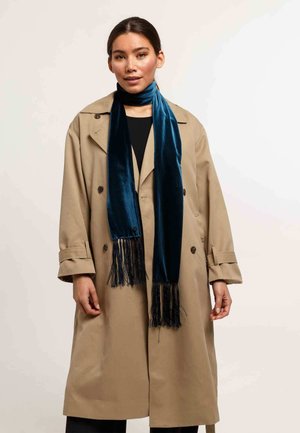 Donna che indossa un trench beige sopra un outfit nero e una lunga sciarpa di velluto blu scuro con frange, in piedi contro uno sfondo neutro.