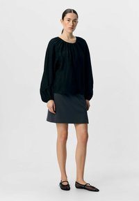 Schwarze strukturierte Bluse mit gerafftem Ausschnitt, kombiniert mit einem dunkelgrauen Rock. Die Schuhe bestehen aus schwarzem Netzmaterial mit Schleifenakzenten.