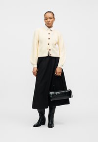 Γυναίκα με πλεξούδες, που φοράει κρεμ ζακέτα με κουμπιά, μαύρη midi φούστα, μαύρες μπότες και κρατάει μαύρη ορθογώνια τσάντα, μπροστά σε λευκό φόντο.