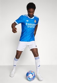 Blue soccer jersey with white accents, "Emirates" logo, paired with white shorts and blue-striped socks. A colorful soccer ball is at his feet.