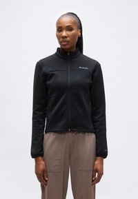 Γυναίκα με σκούρα κοτσίδα που φοράει μαύρο μπουφάν Columbia με φερμουάρ και παντελόνι σε απόχρωση τάουπε, στέκεται μπροστά σε ένα απλό ανοιχτό φόντο.