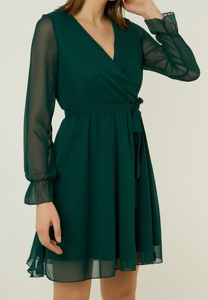 Kobieta w ciemnozielonej, długorękawowej sukience z szyfonu z kopertowym zapięciem, wiązaną w pasie i z falbankami na mankietach, stojąca na jasnym tle.