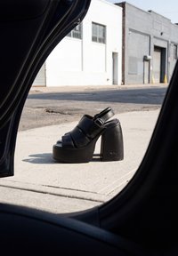 Sandale noire à talon haut et plateforme avec une bride à boucle, posée sur un trottoir en béton, encadrée par une porte de voiture ouverte dans un environnement urbain.